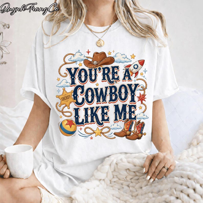 May include: A white t-shirt featuring a graphic print with the text "YOU'RE A COWBOY LIKE ME" in blue and orange. The design includes a brown cowboy hat, a sheriff star badge, a rocket, a colorful ball, and a pair of brown cowboy boots.