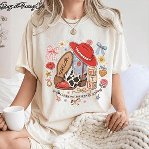 Puede incluir: Una camiseta color crema con un estampado gr&aacute;fico de una bota de vaquero, un sombrero rojo, estampado de vaca y el n&uacute;mero 13. El dise&ntilde;o incluye bloques con las letras T, S, S, una herradura, flores, lazos y el texto I KNEW IT I KNEW YOU.