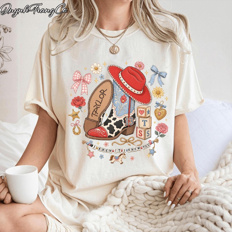 Puede incluir: Una camiseta color crema con un estampado gr&aacute;fico de una bota de vaquero, un sombrero rojo, estampado de vaca y el n&uacute;mero 13. El dise&ntilde;o incluye bloques con las letras T, S, S, una herradura, flores, lazos y el texto I KNEW IT I KNEW YOU.
