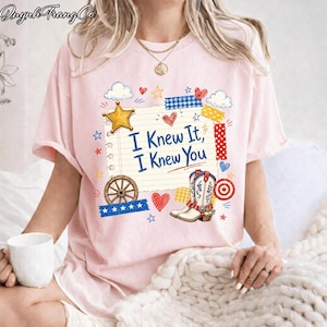 May include: A light pink short-sleeve t-shirt featuring a graphic of a lined notebook page with the text "I Knew It, I Knew You" in blue. The design includes a yellow star, a brown wagon wheel, a cowboy boot, and various red and blue accents.