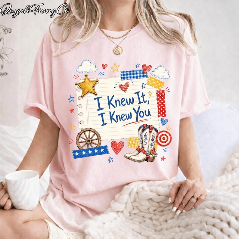 May include: A light pink short-sleeve t-shirt featuring a graphic of a lined notebook page with the text "I Knew It, I Knew You" in blue. The design includes a yellow star, a brown wagon wheel, a cowboy boot, and various red and blue accents.