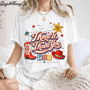 May include: A white short-sleeve t-shirt featuring a graphic print with the text "I Knew It, I Knew You" in red and blue. The design includes a red cowboy boot, a red cowboy hat, a yellow sheriff star, and colorful blocks spelling "TS5".