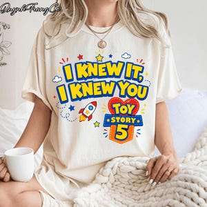 Pu&ograve; includere: Una t-shirt a maniche corte color crema con una stampa grafica che riporta il testo I KNEW IT, I KNEW YOU e TOY STORY 5. Il design include un razzo colorato, piccole stelle e nuvole in blu, rosso e giallo, centrato sul petto.