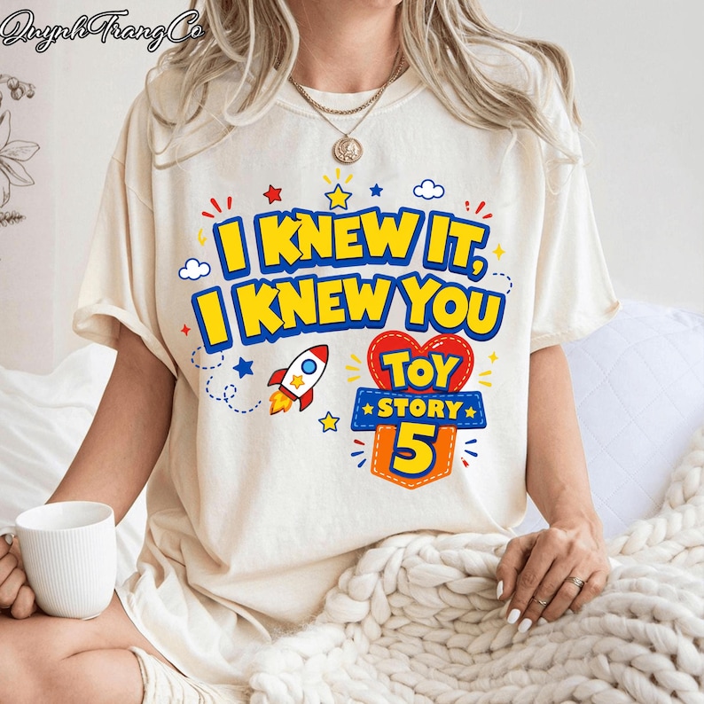 Pu&ograve; includere: Una t-shirt a maniche corte color crema con una stampa grafica che riporta il testo I KNEW IT, I KNEW YOU e TOY STORY 5. Il design include un razzo colorato, piccole stelle e nuvole in blu, rosso e giallo, centrato sul petto.