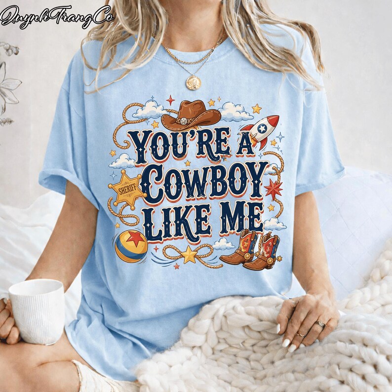 May include: A light blue t-shirt featuring a graphic print with the text "YOU'RE A COWBOY LIKE ME" in dark blue and orange. The design includes a brown cowboy hat, a sheriff star badge, a rocket, a colorful ball, and cowboy boots with rope.