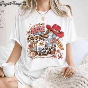 May include: A white short-sleeve t-shirt featuring a graphic print of a cowboy boot with a cow print pattern, a red cowboy hat, three stacked alphabet blocks with the letters T, S, and S, and the text Ride The Adventure, Taylor, and Friends Forever.