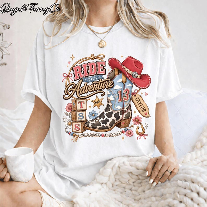 May include: A white short-sleeve t-shirt featuring a graphic print of a cowboy boot with a cow print pattern, a red cowboy hat, three stacked alphabet blocks with the letters T, S, and S, and the text Ride The Adventure, Taylor, and Friends Forever.