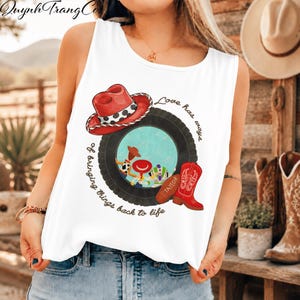 May include: A white sleeveless tank top featuring a graphic print of a red cowboy hat, a black tyre, a red cowboy boot, and cartoon characters. The text reads, "Love has ways of bringing things back to life" and "TAYLOR" on the boot.