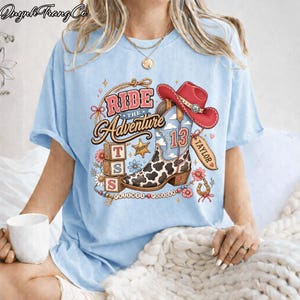 May include: A light blue t-shirt featuring a graphic print of a cow-print cowboy boot, a red cowboy hat, and three stacked blocks with the letters T, S, and S. The text reads Ride The Adventure, Taylor, and Friends Forever with floral accents.