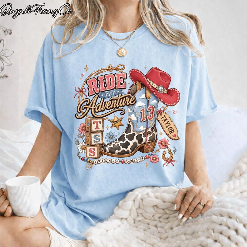 May include: A light blue t-shirt featuring a graphic print of a cow-print cowboy boot, a red cowboy hat, and three stacked blocks with the letters T, S, and S. The text reads Ride The Adventure, Taylor, and Friends Forever with floral accents.