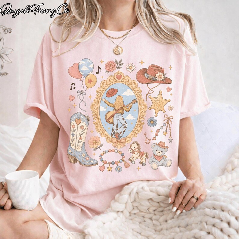 Op de afbeelding: Een lichtroze t-shirt met een centraal ovaal goudkleurig kader met daarin een persoon die gitaar speelt. Rondom het kader staan illustraties van een cowboyhoed, een sheriffster, een gebloemde cowboylaars, ballonnen en een teddybeer.
