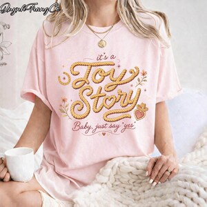 Puede incluir: Una camiseta de manga corta de color rosa claro con el texto "it's a Joy Story" en una fuente estilo cuerda, con la frase "Baby, just say "yes"" debajo. El dise&ntilde;o incluye peque&ntilde;os detalles florales, un coraz&oacute;n y un sombrero vaquero.