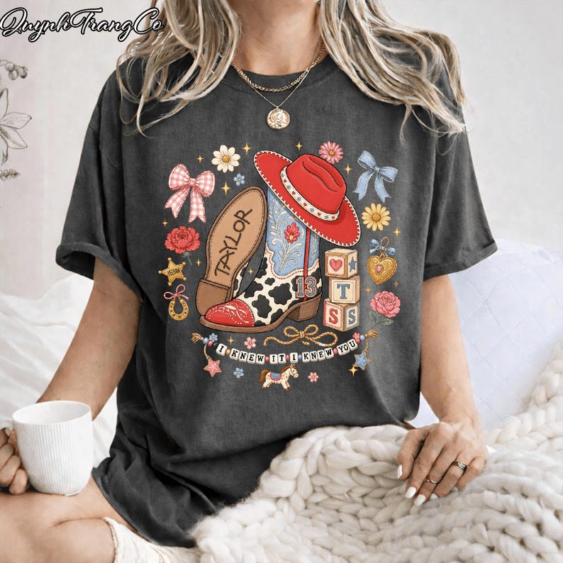 Puede incluir: Una camiseta gris carb&oacute;n con un estampado gr&aacute;fico de un sombrero de vaquero rojo, una bota con estampado de vaca con el nombre Taylor y el n&uacute;mero 13, bloques de letras, lazos, flores y una pancarta con el texto I KNEW IT I KNEW YOU en disposici&oacute;n circular.
