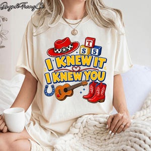 May include: A cream-colored t-shirt featuring a graphic print with a red cowboy hat, alphabet blocks, a guitar, a blue horseshoe, and red cowboy boots. The text reads I KNEW IT I KNEW YOU in bold yellow letters with a blue outline.