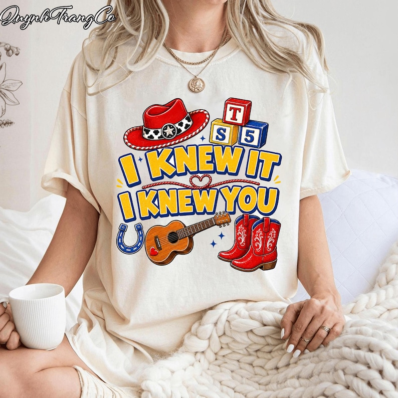 May include: A cream-colored t-shirt featuring a graphic print with a red cowboy hat, alphabet blocks, a guitar, a blue horseshoe, and red cowboy boots. The text reads I KNEW IT I KNEW YOU in bold yellow letters with a blue outline.