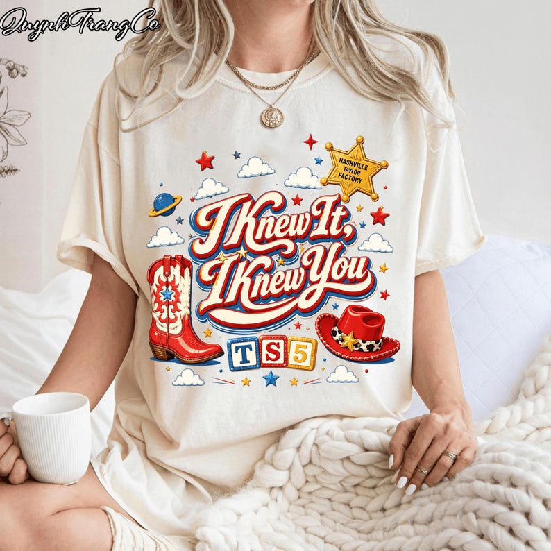 May include: A cream-colored t-shirt featuring a graphic print with the text "I Knew It, I Knew You" in red and blue. The design includes a red cowboy boot, a red cowboy hat, a yellow sheriff star labeled "Nashville Taylor Factory," and "TS5" blocks.