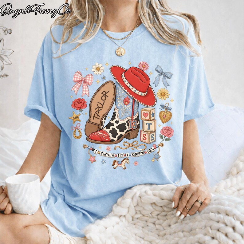 Puede incluir: Una camiseta azul claro con un estampado gr&aacute;fico de una bota de vaquero con estampado de vaca, un sombrero de vaquero rojo, bloques de letras, flores, lazos y una pancarta con el texto I KNEW IT I KNEW YOU. La bota tiene el nombre TAYLOR y el 13.