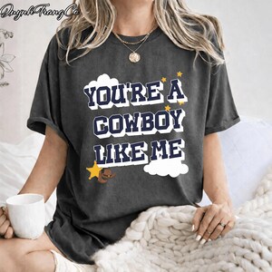 May include: A charcoal gray short-sleeve t-shirt featuring the dark blue text "YOU'RE A COWBOY LIKE ME" in a bold, blocky font. The design includes white cloud shapes, small yellow stars, a yellow sheriff star badge, and a brown cowboy hat graphic.