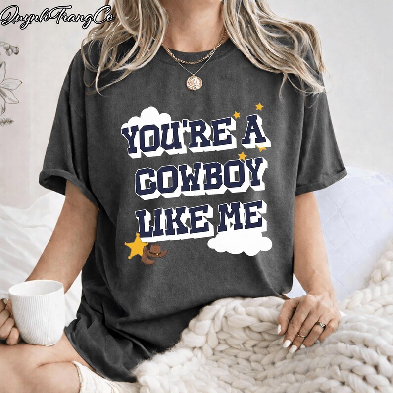 May include: A charcoal gray short-sleeve t-shirt featuring the dark blue text "YOU'RE A COWBOY LIKE ME" in a bold, blocky font. The design includes white cloud shapes, small yellow stars, a yellow sheriff star badge, and a brown cowboy hat graphic.
