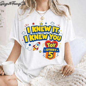 Pu&ograve; includere: Una t-shirt bianca a maniche corte con una stampa grafica colorata con il testo I KNEW IT, I KNEW YOU e TOY STORY 5. Il design include un razzo rosso e bianco, un cuore rosso, stelle gialle e nuvole blu sulla parte anteriore.