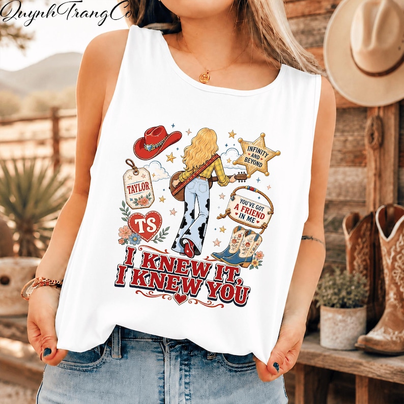 May include: A white sleeveless tank top featuring a graphic print of a blonde person with a guitar, a red cowboy hat, a sheriff star, cowboy boots, and floral accents. The text reads "TAYLOR", "TS", "INFINITY AND BEYOND", "YOU'VE GOT A FRIEND IN ME", and "I KNEW IT, I KNEW YOU".