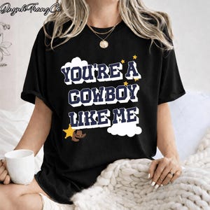 May include: A black short-sleeved t-shirt featuring the white and navy blue text "YOU'RE A COWBOY LIKE ME" in a bold font. The design includes a yellow sheriff star, a brown cowboy hat, and small yellow stars against white cloud shapes.