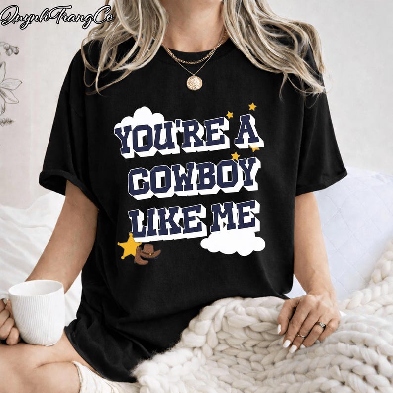 May include: A black short-sleeved t-shirt featuring the white and navy blue text "YOU'RE A COWBOY LIKE ME" in a bold font. The design includes a yellow sheriff star, a brown cowboy hat, and small yellow stars against white cloud shapes.