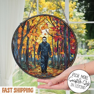 May include: Round stained glass suncatcher featuring a figure in a dark jumpsuit and mask walking through a colorful autumn forest. The artwork is framed in a black metal border and hangs from a chain. The image has the text "Pick More Suncatcher Style in Listing!"
