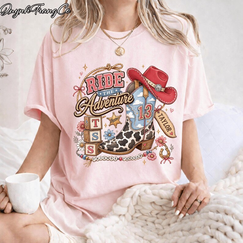 May include: A light pink t-shirt featuring a graphic print of a cow-print cowboy boot with the number 13, a red cowboy hat, three alphabet blocks with the letters T, S, and S, flowers, a lasso, and the text Ride the Adventure and Taylor.