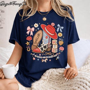 Puede incluir: Una camiseta azul marino con un estampado gr&aacute;fico de una bota vaquera con manchas de vaca, un sombrero vaquero rojo, bloques de alfabeto con las letras T y S, lazos rosas, flores y una pancarta con el texto I KNEW IT I KNEW YOU y el nombre TAYLOR.