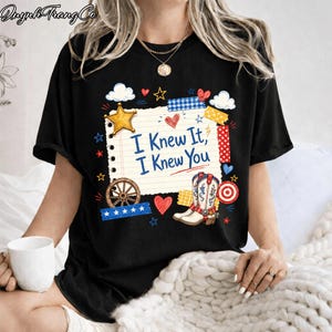 May include: A black t-shirt featuring a central graphic of a lined notepad page with the text "I Knew It, I Knew You" in blue. The design includes a yellow star, a brown wagon wheel, white and red cowboy boots, hearts, and colorful tape strips.