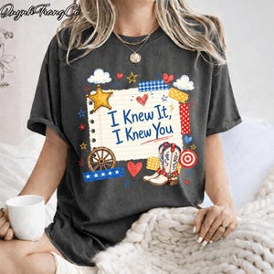 May include: A charcoal gray t-shirt featuring a central graphic of a lined notepad page with the text "I Knew It, I Knew You" in blue. The design includes a yellow star, a wagon wheel, cowboy boots, hearts, and various colorful patterned tape strips.