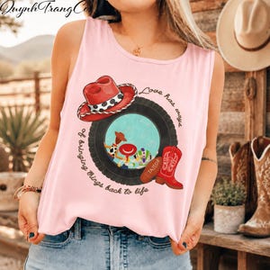May include: A light pink sleeveless tank top featuring a graphic print of a black tyre framing cartoon characters, a red cowboy hat, a red cowboy boot with the name Taylor, and the text Love has ways of bringing things back to life.