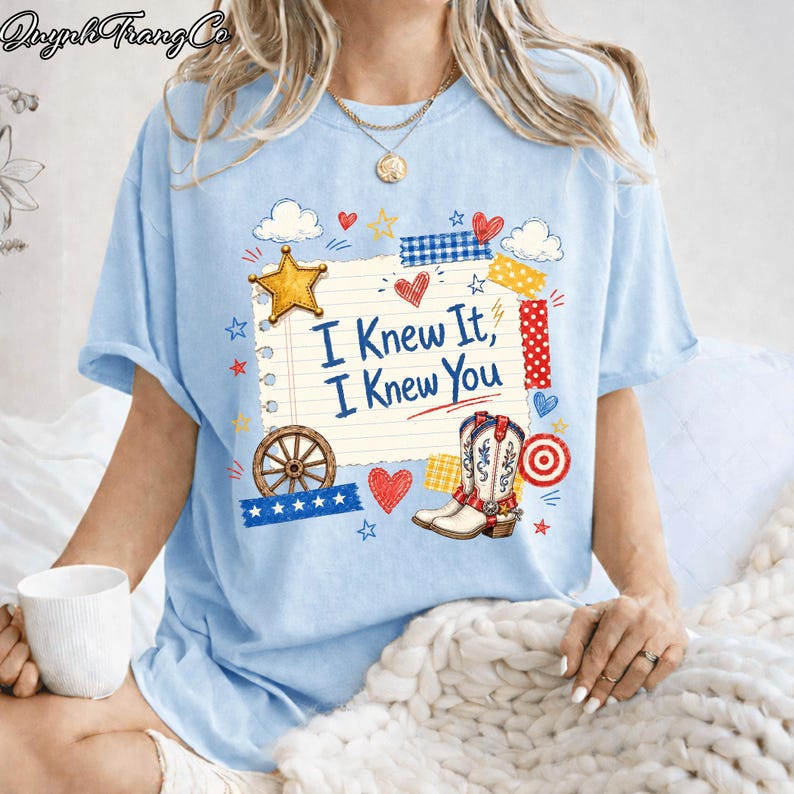 May include: A light blue t-shirt featuring a graphic of a lined notebook page with the text "I Knew It, I Knew You" in blue. The design includes a yellow star, a brown wagon wheel, a pair of western boots, red hearts, and colorful tape accents.