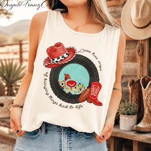 May include: A white sleeveless tank top featuring a graphic print of a red cowboy hat, a black tyre, and a red cowboy boot. The design includes the text "Love has ways of bringing things back to life" and small cartoon characters inside the tyre.