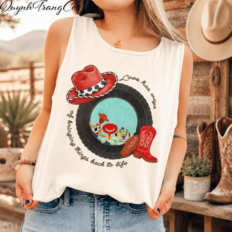 May include: A white sleeveless tank top featuring a graphic print of a red cowboy hat, a black tyre, and a red cowboy boot. The design includes the text "Love has ways of bringing things back to life" and small cartoon characters inside the tyre.