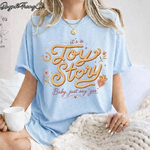 Puede incluir: Una camiseta azul claro con un dise&ntilde;o gr&aacute;fico que incluye el texto "it's a Joy Story" y "Baby, just say 'yes'" en una fuente estilo cuerda. El dise&ntilde;o presenta peque&ntilde;as ilustraciones florales, una herradura y un icono de sombrero vaquero.