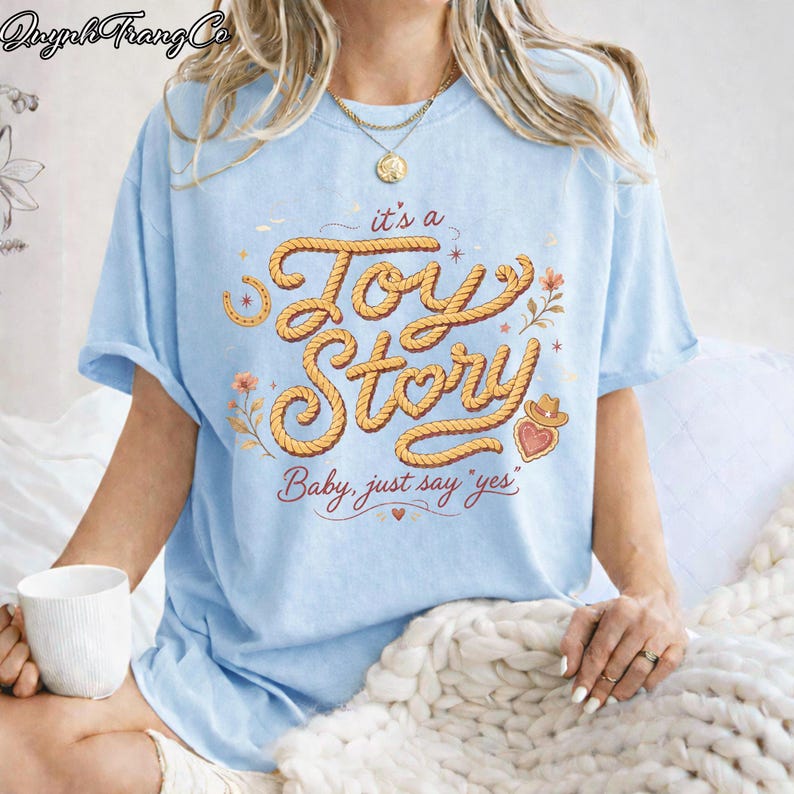 Puede incluir: Una camiseta azul claro con un dise&ntilde;o gr&aacute;fico que incluye el texto "it's a Joy Story" y "Baby, just say 'yes'" en una fuente estilo cuerda. El dise&ntilde;o presenta peque&ntilde;as ilustraciones florales, una herradura y un icono de sombrero vaquero.
