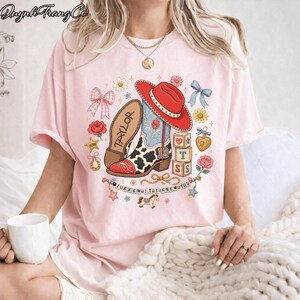 Puede incluir: Una camiseta rosa claro con un estampado gr&aacute;fico de una bota vaquera con un sombrero rojo, un patr&oacute;n de manchas de vaca y el nombre Taylor. El dise&ntilde;o incluye bloques de letras, lazos, flores, un coraz&oacute;n y el texto I KNEW IT I KNEW YOU.