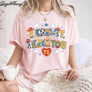 Puede incluir: Una camiseta de manga corta de color rosa claro con un estampado gr&aacute;fico colorido con el texto I KNEW IT, I KNEW YOU y TS dentro de un coraz&oacute;n rojo. El dise&ntilde;o incluye sombreros de vaquero, botas, cactus, estrellas, nubes y motivos de cuerda.