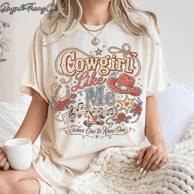 May include: A cream-coloured t-shirt featuring a graphic print with the text Cowgirl Like Me Takes One to Know One. The design includes a lasso, a cowboy hat, a horseshoe, a rocking horse, musical notes, stars, and red roses in a vintage style.