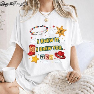 May include: A white short-sleeve t-shirt featuring a colorful graphic print with red cowboy boots, a red cowboy hat, a beaded bracelet, a yellow star badge with the text "INFINITY AND BEYOND," and the text "I KNEW IT, I KNEW YOU, TS5."