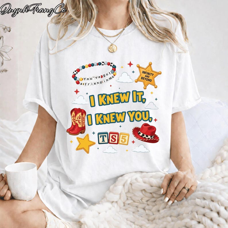 May include: A white short-sleeve t-shirt featuring a colorful graphic print with red cowboy boots, a red cowboy hat, a beaded bracelet, a yellow star badge with the text "INFINITY AND BEYOND," and the text "I KNEW IT, I KNEW YOU, TS5."