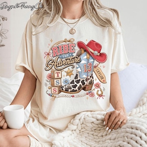 May include: A cream-colored t-shirt featuring a graphic print of a cowboy boot with a cow print pattern, a red cowboy hat, three stacked blocks with the letters T, S, and S, a star, and the text Ride The Adventure, Taylor, and Friends Forever.