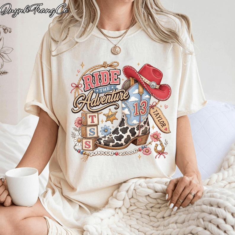 May include: A cream-colored t-shirt featuring a graphic print of a cowboy boot with a cow print pattern, a red cowboy hat, three stacked blocks with the letters T, S, and S, a star, and the text Ride The Adventure, Taylor, and Friends Forever.