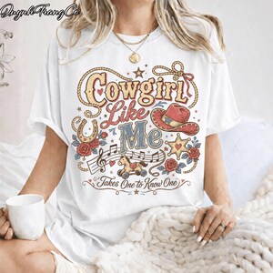 May include: A white short-sleeve t-shirt featuring a graphic print with the text Cowgirl Like Me and Takes One to Know One. The design includes a red cowboy hat, a horseshoe, a rocking horse, musical notes, red roses, and a sheriff star.