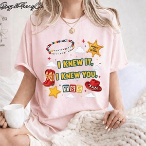 May include: A light pink t-shirt featuring a graphic print with a beaded bracelet, a yellow sheriff star badge with the text "INFINITY AND BEYOND", red cowboy boots, a red cowboy hat, and blocks spelling "TS5" with the text "I KNEW IT, I KNEW YOU".