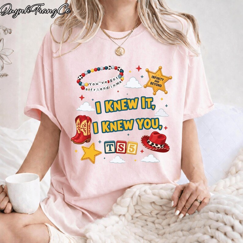 May include: A light pink t-shirt featuring a graphic print with a beaded bracelet, a yellow sheriff star badge with the text "INFINITY AND BEYOND", red cowboy boots, a red cowboy hat, and blocks spelling "TS5" with the text "I KNEW IT, I KNEW YOU".