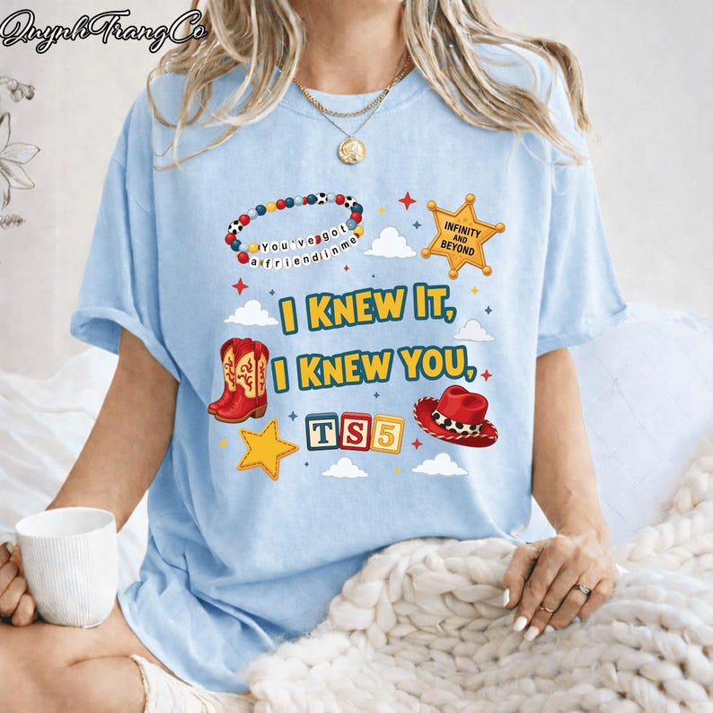 May include: A light blue t-shirt featuring a graphic print with the text I KNEW IT, I KNEW YOU, TS5, INFINITY AND BEYOND, and YOU'VE GOT A FRIEND IN ME. The design includes a beaded bracelet, a gold sheriff star, red cowboy boots, and a red hat.