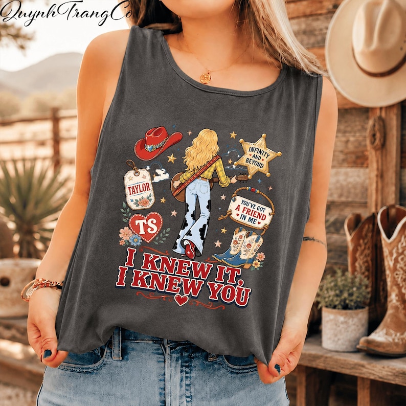 May include: A charcoal grey sleeveless tank top featuring a graphic print of a person with blonde hair playing a guitar, wearing cow print trousers. The design includes a red cowboy hat, a sheriff star, cowboy boots, and the text I KNEW IT, I KNEW YOU.